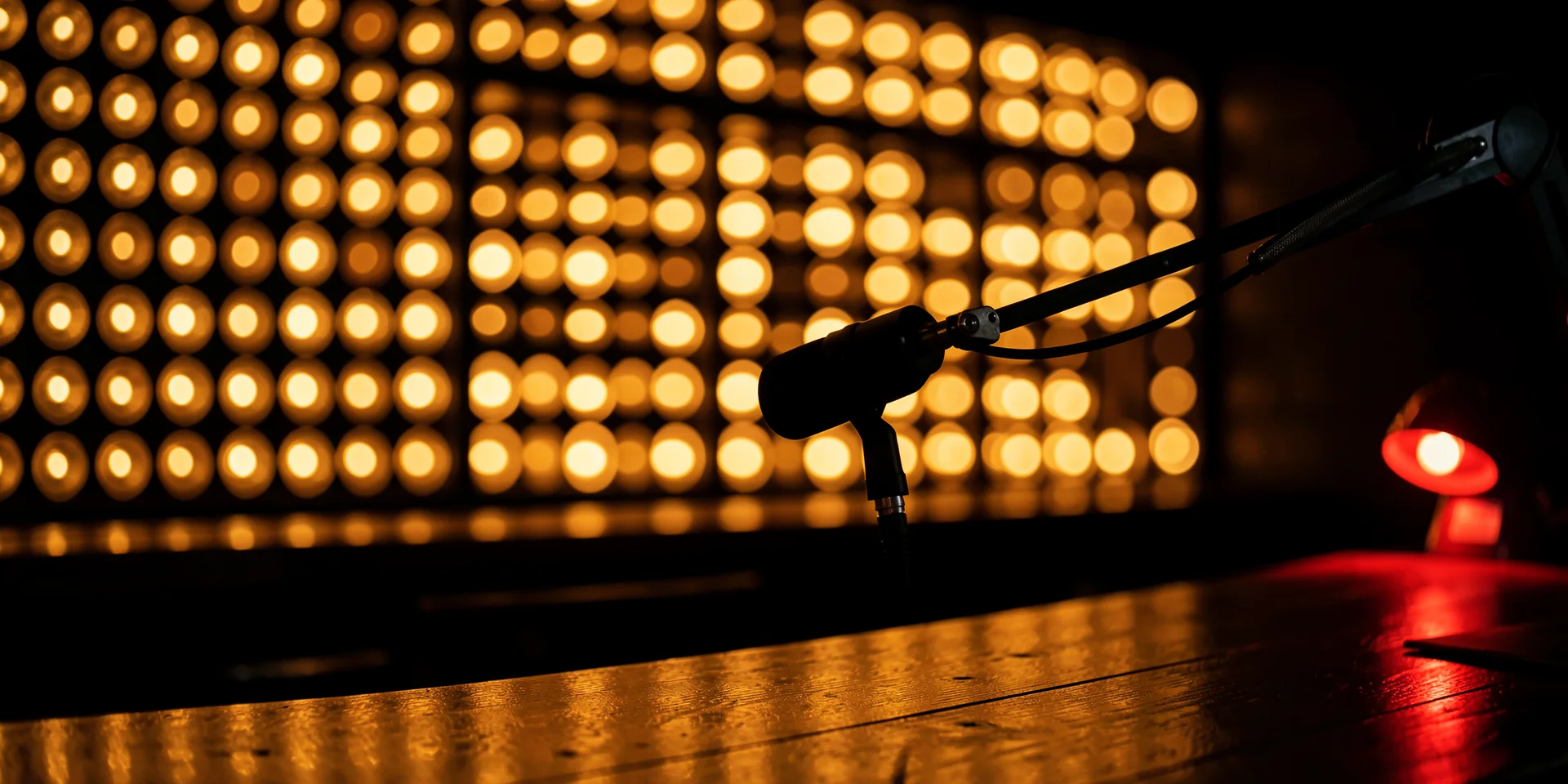 A podcast microphone on a dark bar in front of a wall of glowing amber scoreboard bulbs, with a red recording light.